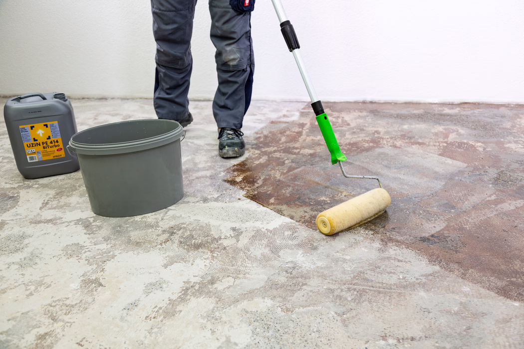 UZIN_Application Image_PE 414_9406_2018-11 A worker's lower legs and feet are visible as they apply a brown primer or adhesive to a bare concrete floor with a long-handled paint roller. A gray plastic bucket and a gray canister labeled "UZIN PE 414 Biturbo" stand nearby against a white wall. The worker wears gray work trousers and black safety shoes; the floor shows patches of old adhesive and ground concrete while the roller spreads the liquid evenly to prepare the subfloor.